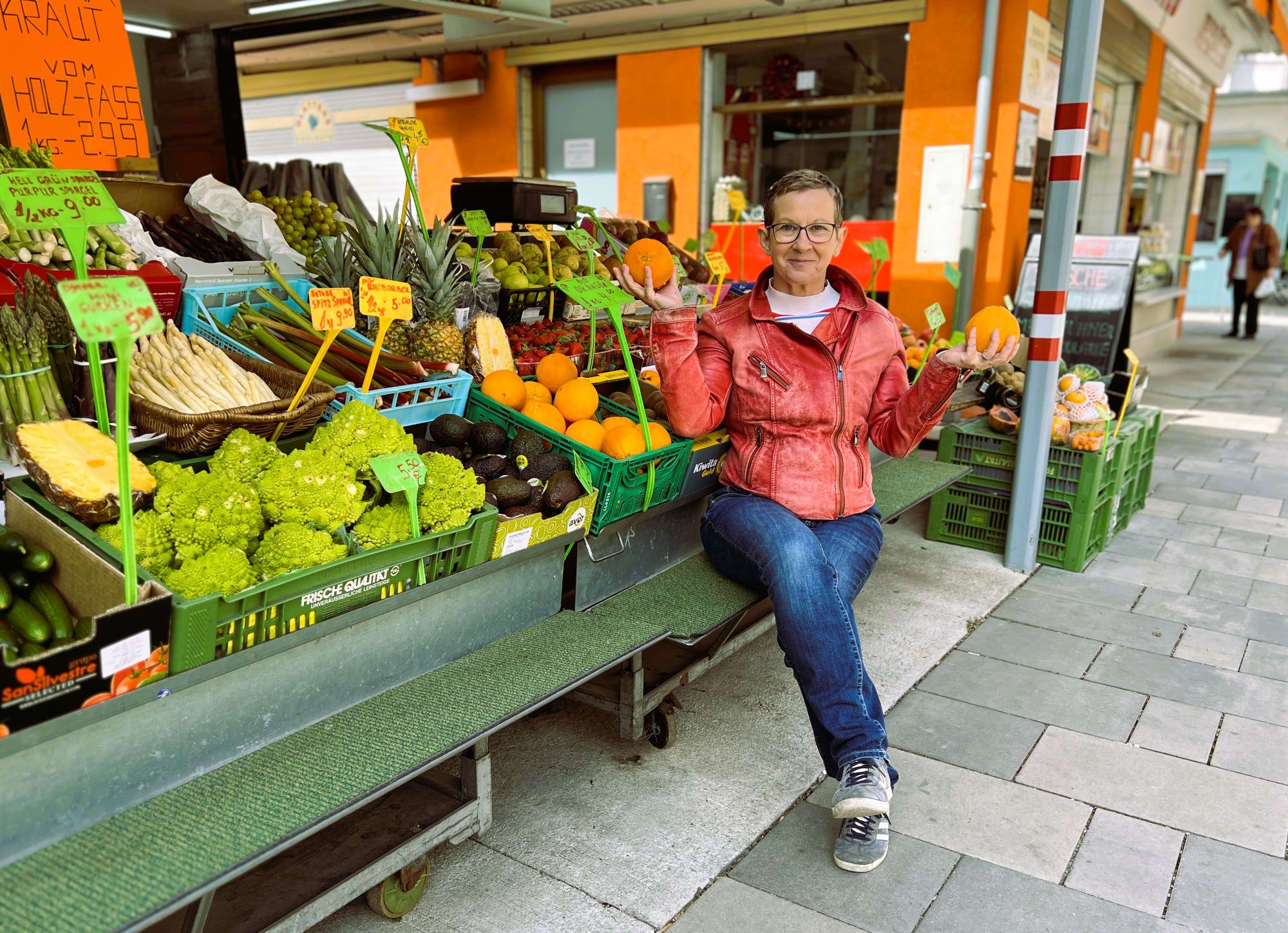 Eine Frau sitzt bei einem Gemüsestand am Markt und hat zwei Orangen in den Händen